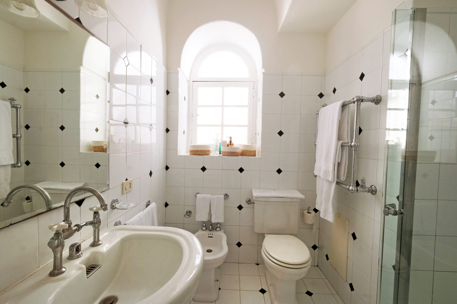 Modern bathroom featuring white tiled walls with black accents, a glass shower, a bidet, and a vintage-style sink with dual faucets. Natural light streams through a window, illuminating the space. Fresh towels are neatly arranged, enhancing the clean and inviting atmosphere. | Only on SopranoVillas