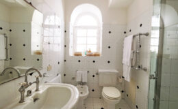 Modern bathroom featuring white tiled walls with black accents, a glass shower, a bidet, and a vintage-style sink with dual faucets. Natural light streams through a window, illuminating the space. Fresh towels are neatly arranged, enhancing the clean and inviting atmosphere.