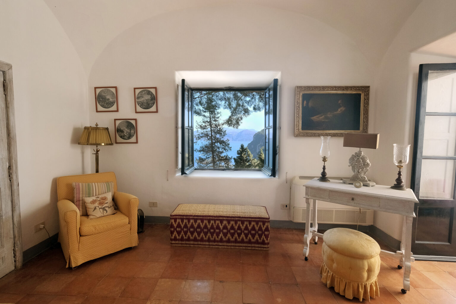 Cozy living room with a yellow armchair, decorative lamp, and a vintage table, featuring a stunning view of mountains and trees through an open window. The room has terracotta tile flooring and framed artwork on the walls. | Only on SopranoVillas
