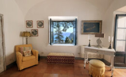 Cozy living room with a yellow armchair, decorative lamp, and a vintage table, featuring a stunning view of mountains and trees through an open window. The room has terracotta tile flooring and framed artwork on the walls.