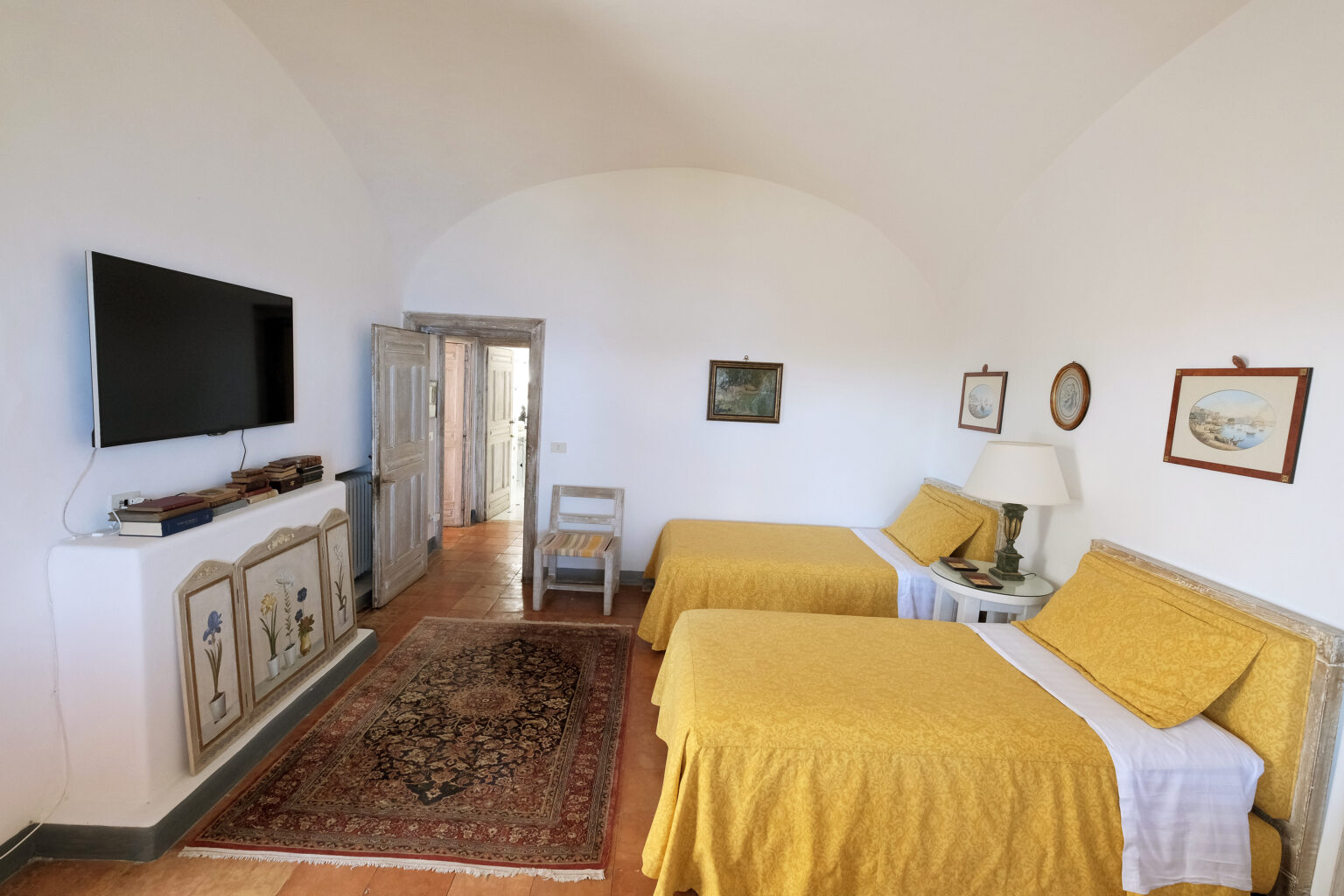 Cozy guest room featuring two twin beds with yellow bedspreads, a flat-screen TV mounted on the wall, a decorative area rug, and artwork hanging on the walls. The room includes a wooden chair and a door leading to a hallway, creating a warm and inviting atmosphere. Ideal for travelers seeking comfort and style. | Only on SopranoVillas