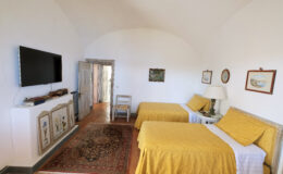 Cozy guest room featuring two twin beds with yellow bedspreads, a flat-screen TV mounted on the wall, a decorative area rug, and artwork hanging on the walls. The room includes a wooden chair and a door leading to a hallway, creating a warm and inviting atmosphere. Ideal for travelers seeking comfort and style.