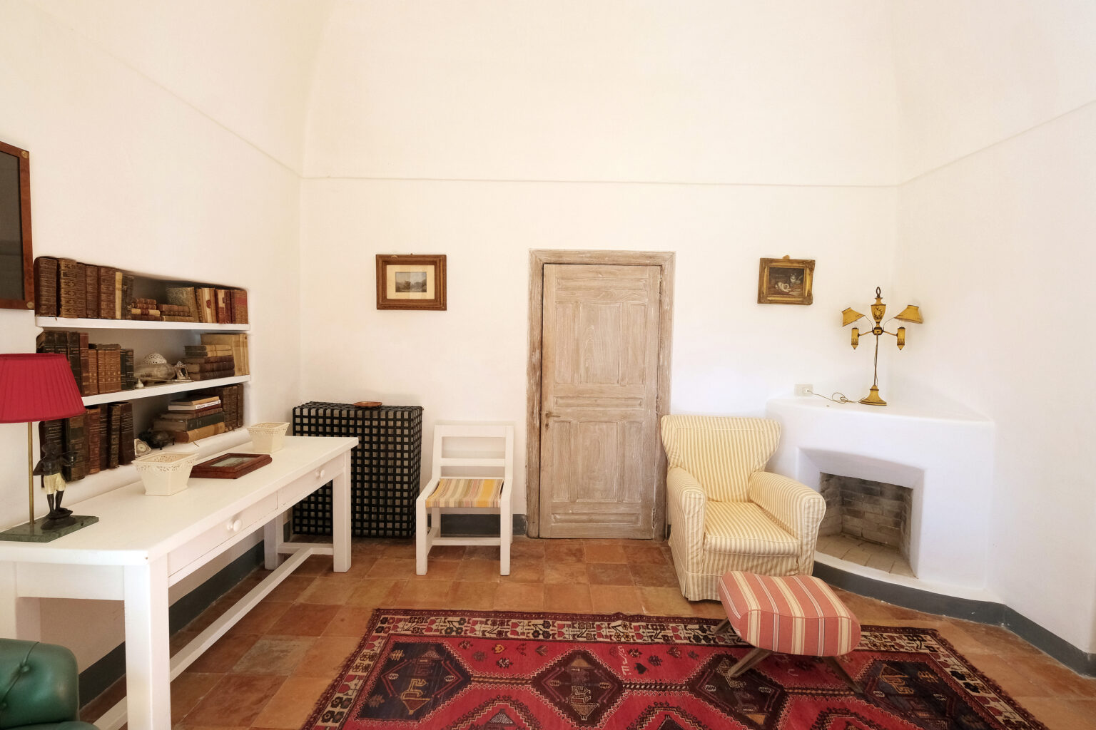 Cozy interior of a minimalist room featuring a white desk, a vintage chair, a comfortable armchair, and decorative elements including a lamp and framed artwork. The space has a warm atmosphere with a patterned rug and a rustic door. | Only on SopranoVillas