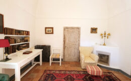 Cozy interior of a minimalist room featuring a white desk, a vintage chair, a comfortable armchair, and decorative elements including a lamp and framed artwork. The space has a warm atmosphere with a patterned rug and a rustic door.