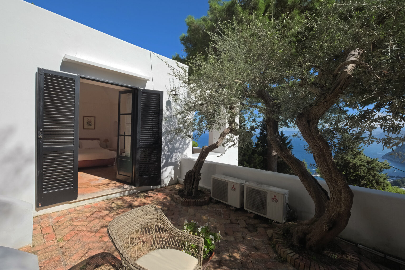 Cozy outdoor view of a modern villa featuring an olive tree, open black shutters, and a serene landscape. The image showcases a brick patio and air conditioning units, highlighting a blend of nature and contemporary design. | Only on SopranoVillas