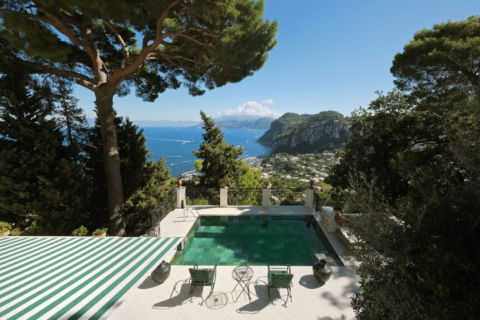 Scenic view of a luxury villa with a swimming pool overlooking the coastline of Capri, surrounded by lush greenery and mountains under a clear blue sky. | Only on SopranoVillas