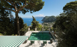 Scenic view of a luxury villa with a swimming pool overlooking the coastline of Capri, surrounded by lush greenery and mountains under a clear blue sky.