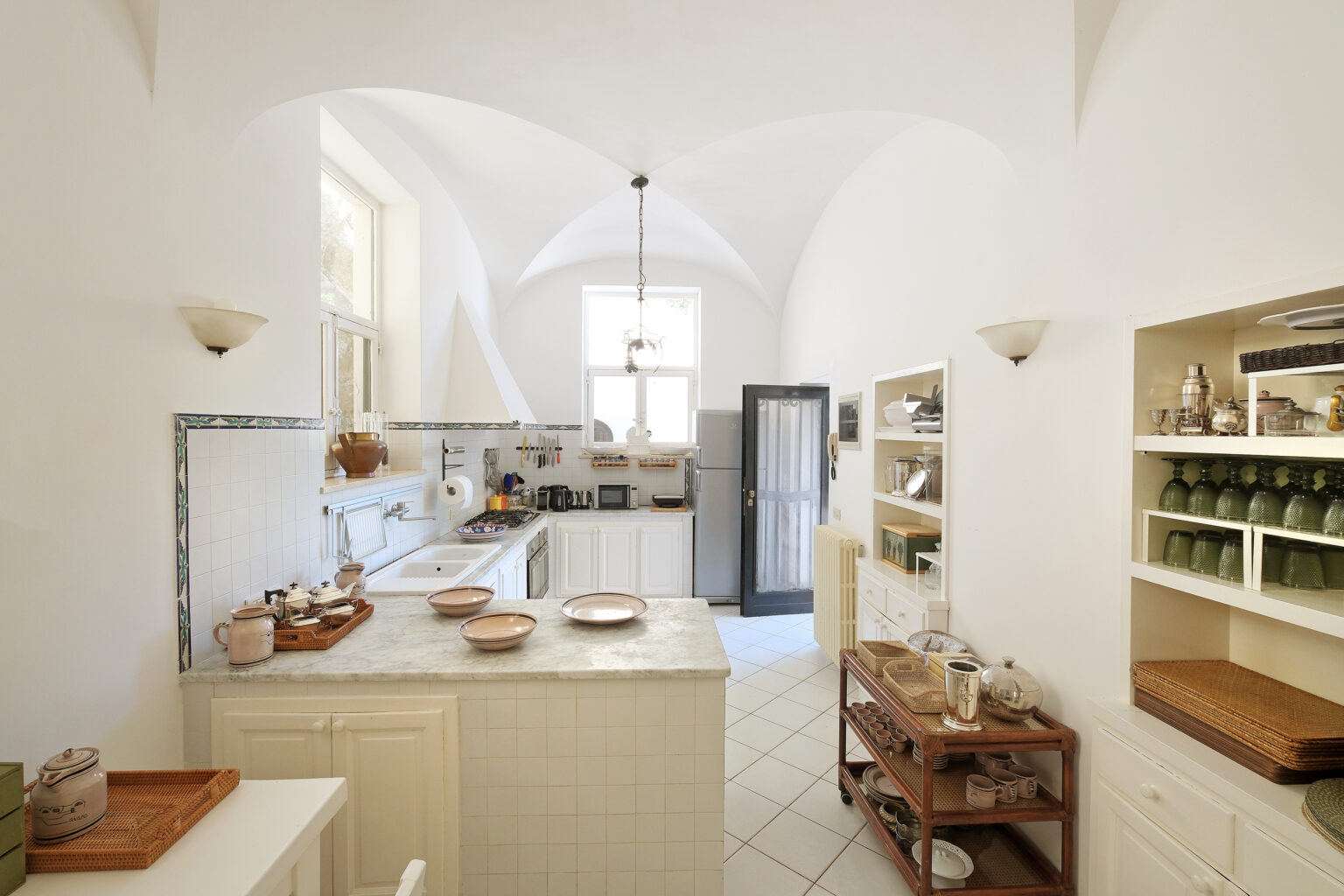 Bright and spacious modern kitchen featuring white cabinetry, a marble countertop, and a unique arched ceiling. The room includes wooden shelving with neatly organized kitchenware and a cozy dining area. Natural light floods the space, enhancing its inviting atmosphere. | Only on SopranoVillas