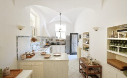Bright and spacious modern kitchen featuring white cabinetry, a marble countertop, and a unique arched ceiling. The room includes wooden shelving with neatly organized kitchenware and a cozy dining area. Natural light floods the space, enhancing its inviting atmosphere.