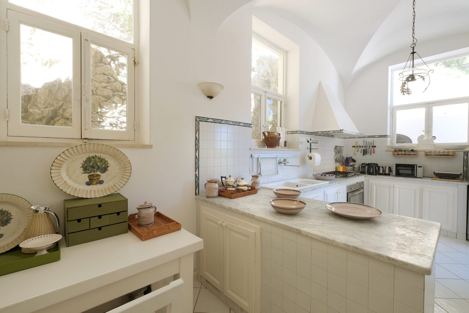 Bright and airy modern kitchen featuring white cabinetry, marble countertops, and natural light streaming through large windows, with decorative dishware and kitchen accessories enhancing the aesthetic. | Only on SopranoVillas