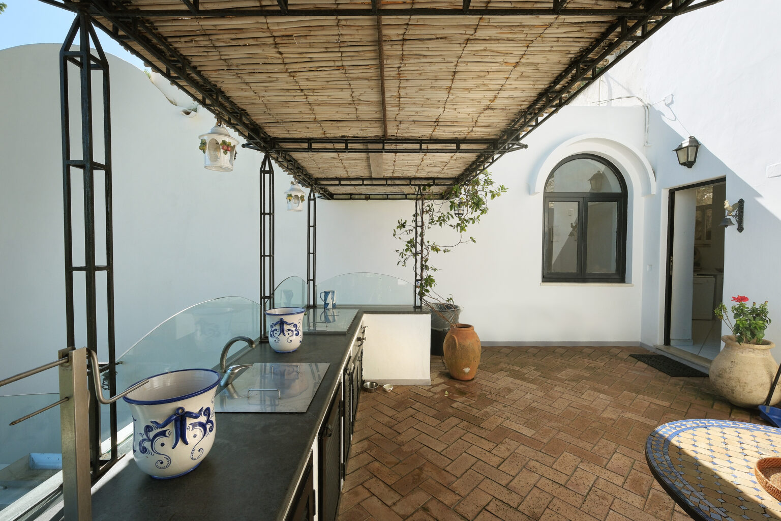 Modern outdoor kitchen area featuring a bamboo roof, decorative pottery, and a tiled table, set in a sunlit courtyard with a whitewashed wall and lush greenery. | Only on SopranoVillas