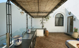 Modern outdoor kitchen area featuring a bamboo roof, decorative pottery, and a tiled table, set in a sunlit courtyard with a whitewashed wall and lush greenery.