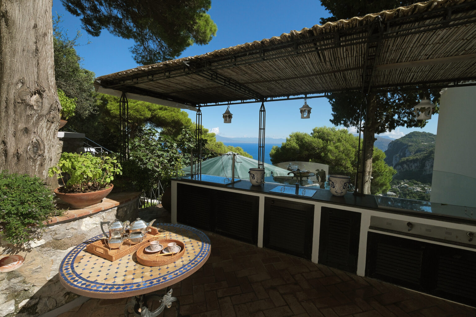 Scenic outdoor kitchen with a stunning sea view, featuring a tiled table set for dining, lush greenery, and a shaded patio area. | Only on SopranoVillas