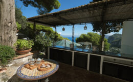 Scenic outdoor kitchen with a stunning sea view, featuring a tiled table set for dining, lush greenery, and a shaded patio area.