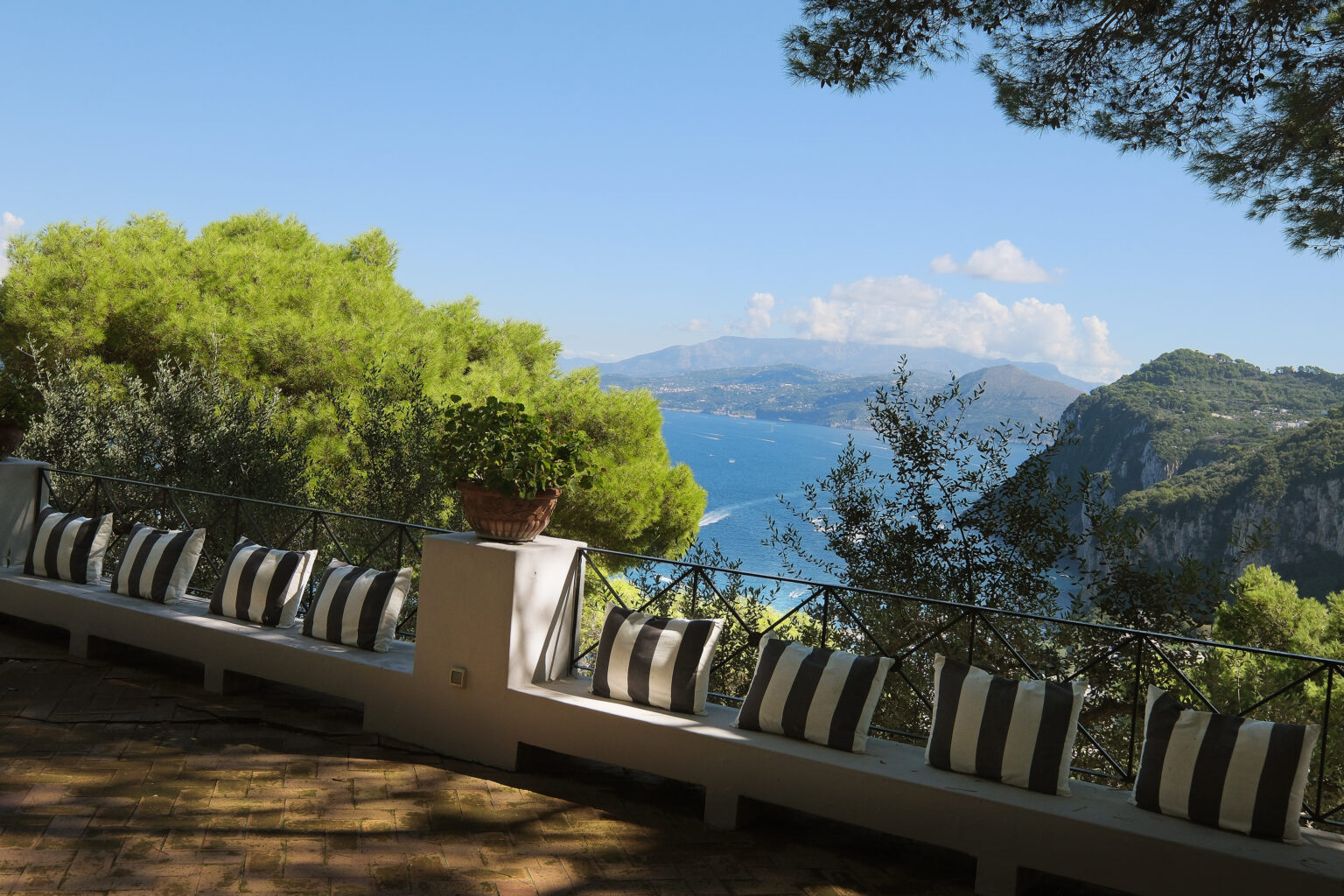 Scenic view from a terrace overlooking the coastline, featuring lush greenery and a row of striped cushions, with mountains and a clear blue sky in the background. | Only on SopranoVillas