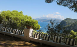 Scenic view from a terrace overlooking the coastline, featuring lush greenery and a row of striped cushions, with mountains and a clear blue sky in the background.