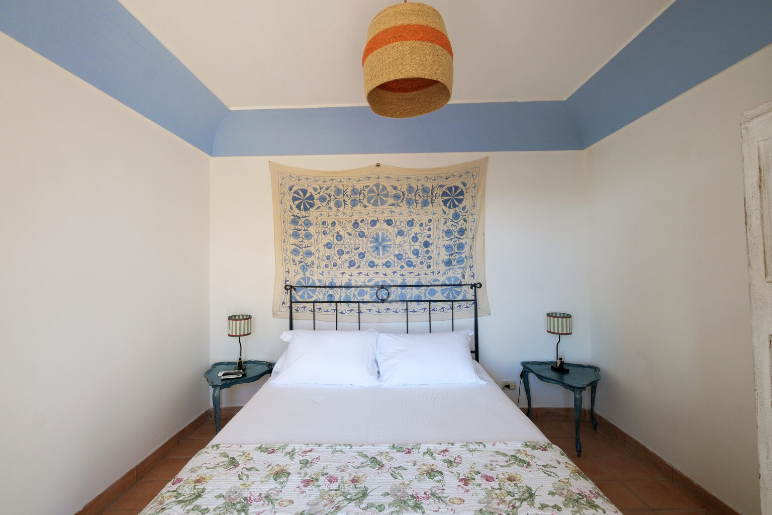 Cozy bedroom featuring a double bed with floral bedding, two bedside tables with lamps, and a decorative wall hanging. The room has light-colored walls and a blue ceiling, creating a serene atmosphere. | Only on SopranoVillas