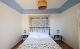 Cozy bedroom featuring a double bed with floral bedding, two bedside tables with lamps, and a decorative wall hanging. The room has light-colored walls and a blue ceiling, creating a serene atmosphere.