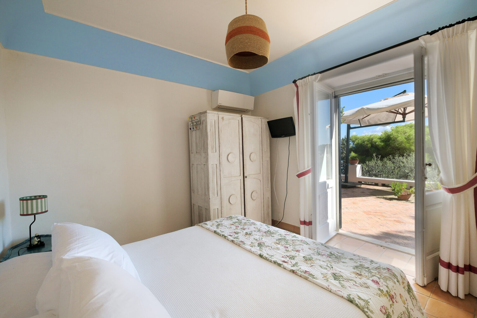 Cozy bedroom featuring a double bed with floral bedding, a wooden wardrobe, and a stylish pendant light. Large glass doors lead to a private patio with outdoor seating and garden views, creating a serene atmosphere. | Only on SopranoVillas