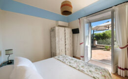Cozy bedroom featuring a double bed with floral bedding, a wooden wardrobe, and a stylish pendant light. Large glass doors lead to a private patio with outdoor seating and garden views, creating a serene atmosphere.