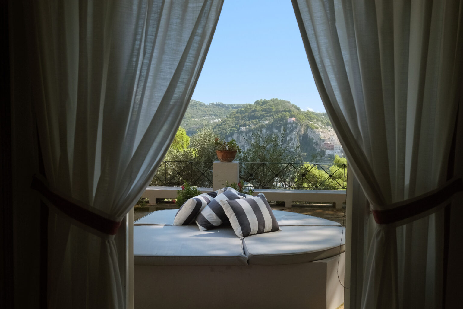 Cozy outdoor seating area with striped cushions, framed by sheer curtains, offering a scenic view of lush hills and clear blue sky. Perfect for relaxation and enjoying nature. | Only on SopranoVillas