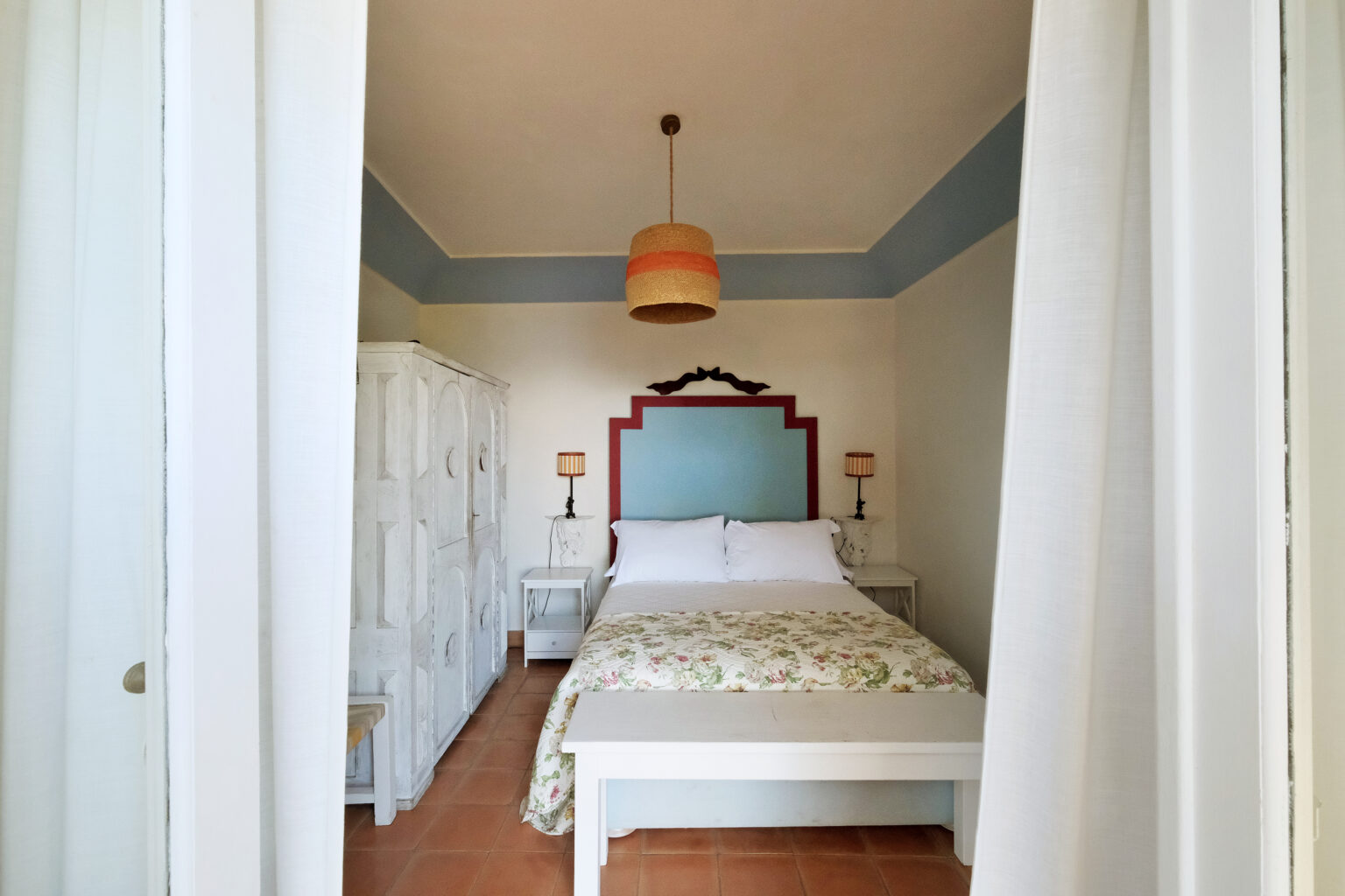Cozy bedroom interior featuring a queen-sized bed with floral bedding, white furniture, and a decorative blue and red accent wall. Soft lighting from a pendant lamp enhances the inviting atmosphere. | Only on SopranoVillas