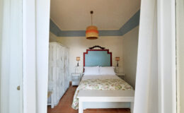 Cozy bedroom interior featuring a queen-sized bed with floral bedding, white furniture, and a decorative blue and red accent wall. Soft lighting from a pendant lamp enhances the inviting atmosphere.