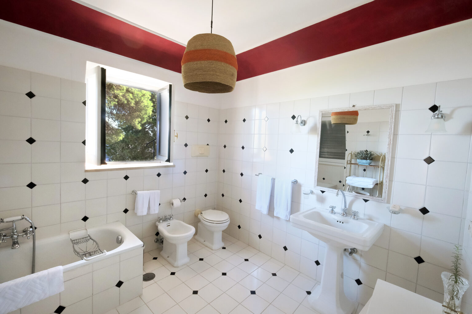 Bright and spacious bathroom featuring a bathtub, toilet, bidet, and sink with a large mirror. The room has white tiled walls with black accents and a red ceiling. Natural light floods in through a window, enhancing the modern aesthetic. | Only on SopranoVillas