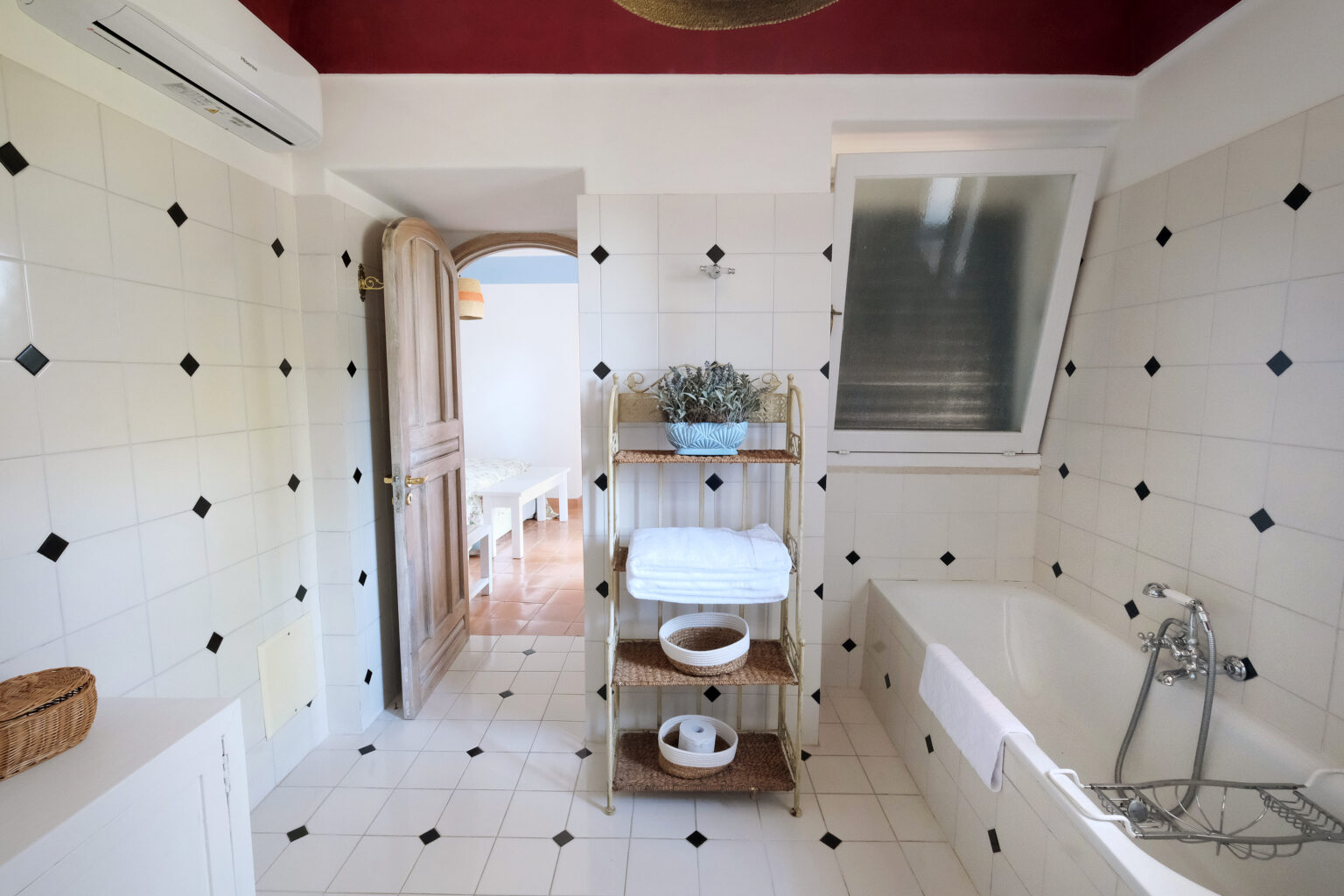 Bright and spacious bathroom featuring white tiled walls with black accents, a bathtub, and a wooden shelf with neatly stacked towels. A door leads to a well-lit area, enhancing the open feel of the space. | Only on SopranoVillas