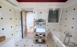 Bright and spacious bathroom featuring white tiled walls with black accents, a bathtub, and a wooden shelf with neatly stacked towels. A door leads to a well-lit area, enhancing the open feel of the space.