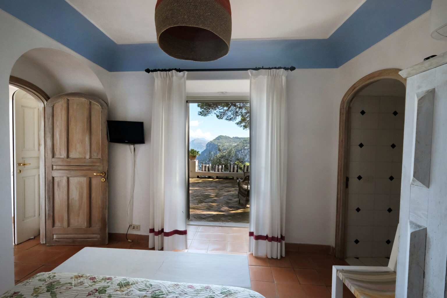 Cozy interior of a room featuring wooden doors, white curtains, and a scenic view of mountains and trees through an open doorway, with a light fixture hanging from the ceiling. | Only on SopranoVillas