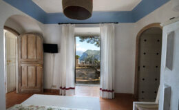 Cozy interior of a room featuring wooden doors, white curtains, and a scenic view of mountains and trees through an open doorway, with a light fixture hanging from the ceiling.