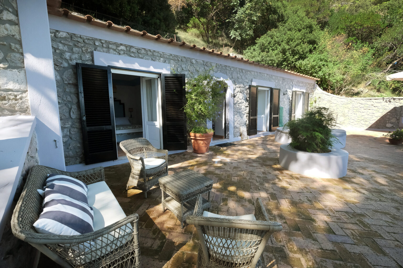 Cozy outdoor seating area with wicker chairs and decorative cushions, featuring a stone cottage surrounded by greenery and large windows, ideal for relaxation in a tranquil setting. | Only on SopranoVillas
