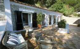 Cozy outdoor seating area with wicker chairs and decorative cushions, featuring a stone cottage surrounded by greenery and large windows, ideal for relaxation in a tranquil setting.