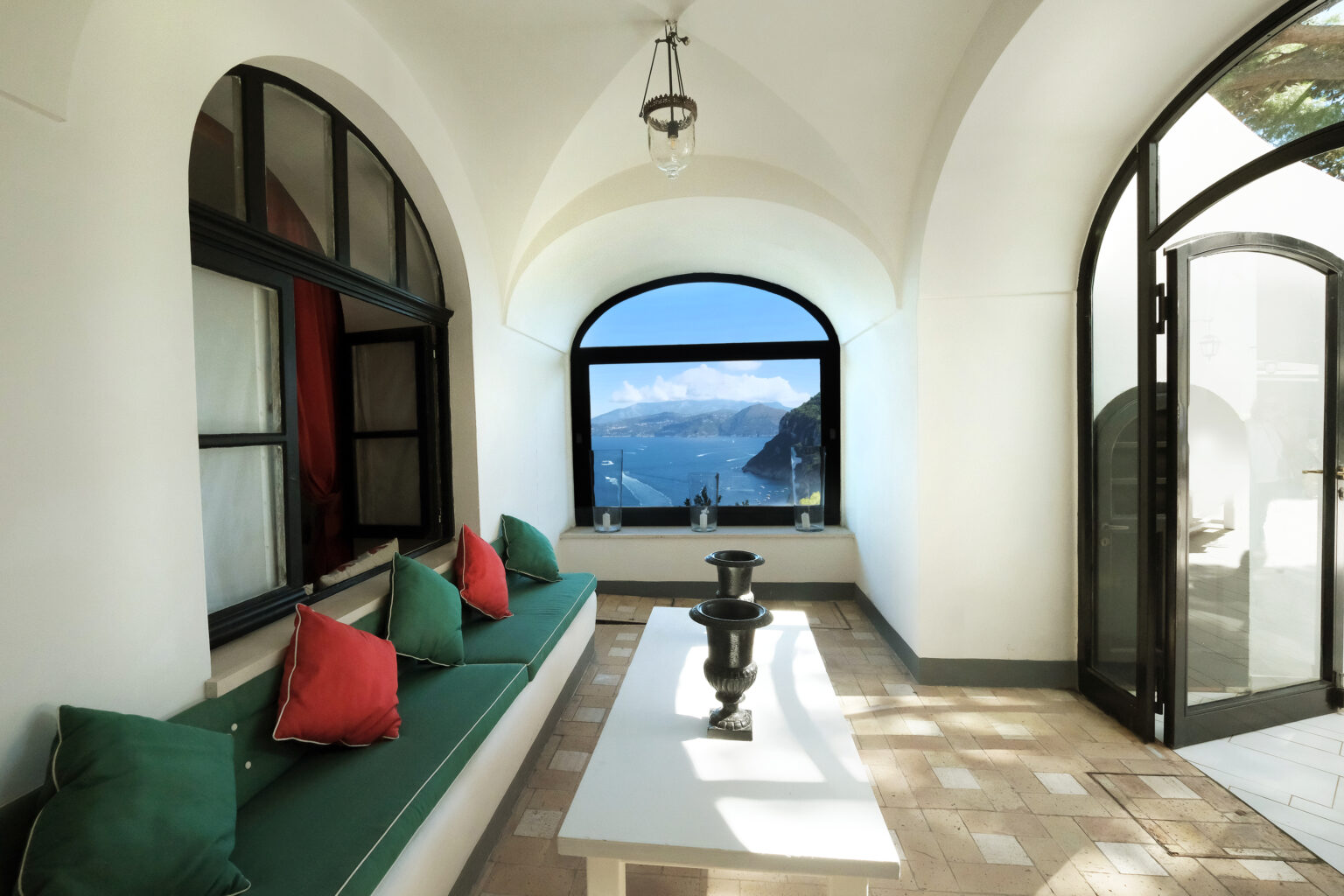 Modern coastal living room with arched windows offering a stunning ocean view, featuring a minimalist design, white walls, and a comfortable seating area adorned with colorful cushions. | Only on SopranoVillas