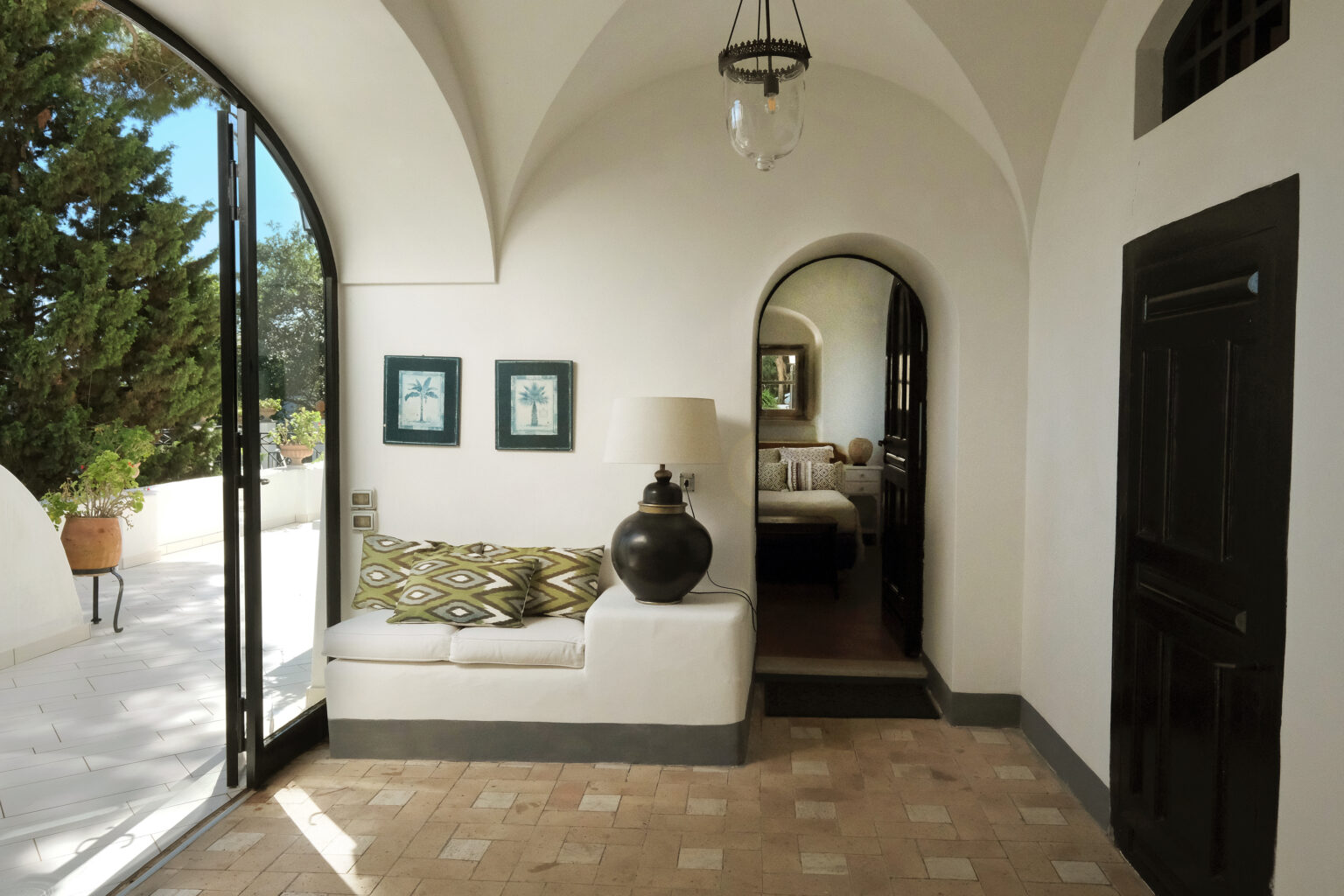 Bright and airy interior of a modern Mediterranean-style home featuring a cozy seating area with patterned cushions, a decorative lamp, and artwork on the walls. Large arched windows provide natural light and a view of the outdoor greenery. A doorway leads to a bedroom area, enhancing the inviting atmosphere. | Only on SopranoVillas