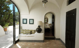 Bright and airy interior of a modern Mediterranean-style home featuring a cozy seating area with patterned cushions, a decorative lamp, and artwork on the walls. Large arched windows provide natural light and a view of the outdoor greenery. A doorway leads to a bedroom area, enhancing the inviting atmosphere.