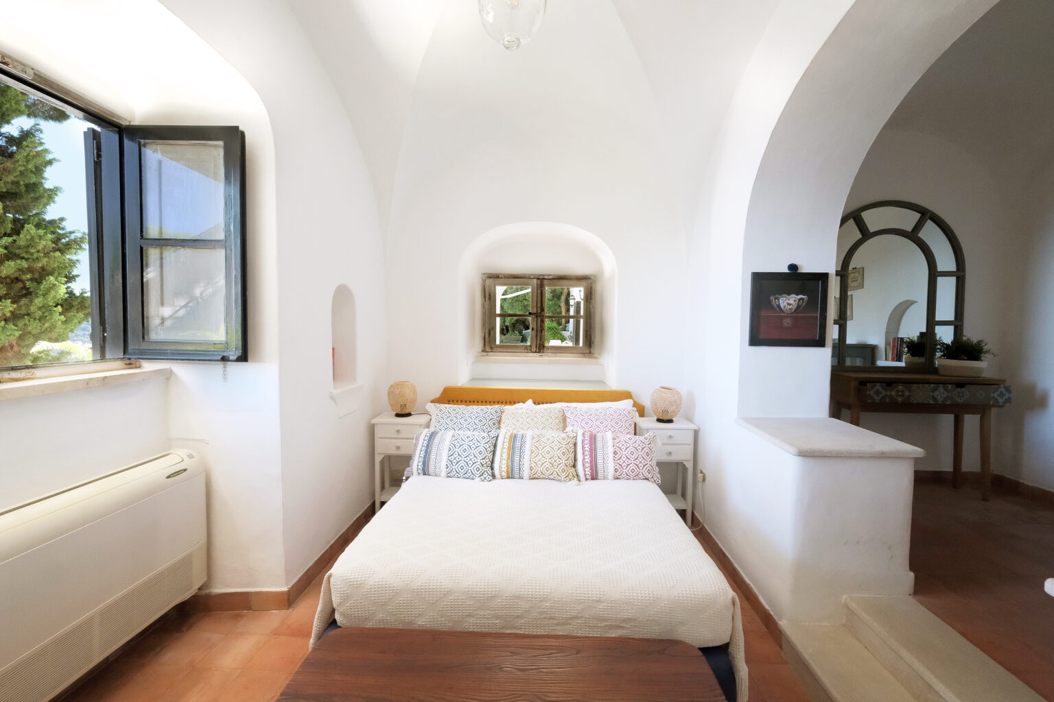 Cozy bedroom with a large bed adorned with colorful pillows, featuring a window that offers natural light and views of greenery. The room has white walls, terracotta tile flooring, and minimalist decor, creating a serene and inviting atmosphere. | Only on SopranoVillas