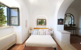 Cozy bedroom with a large bed adorned with colorful pillows, featuring a window that offers natural light and views of greenery. The room has white walls, terracotta tile flooring, and minimalist decor, creating a serene and inviting atmosphere.