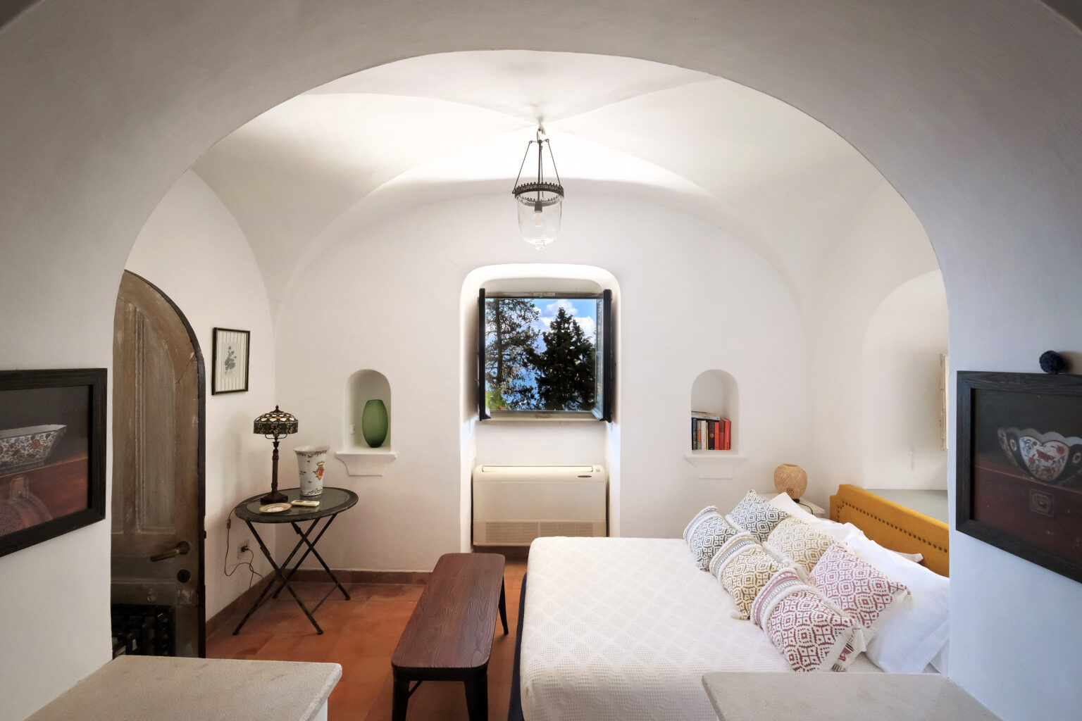 Cozy bedroom with arched ceiling, featuring a double bed adorned with decorative pillows, a wooden coffee table, and a vintage lamp. A window offers a view of greenery, while minimalist decor elements enhance the serene ambiance. | Only on SopranoVillas