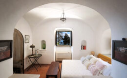 Cozy bedroom with arched ceiling, featuring a double bed adorned with decorative pillows, a wooden coffee table, and a vintage lamp. A window offers a view of greenery, while minimalist decor elements enhance the serene ambiance.