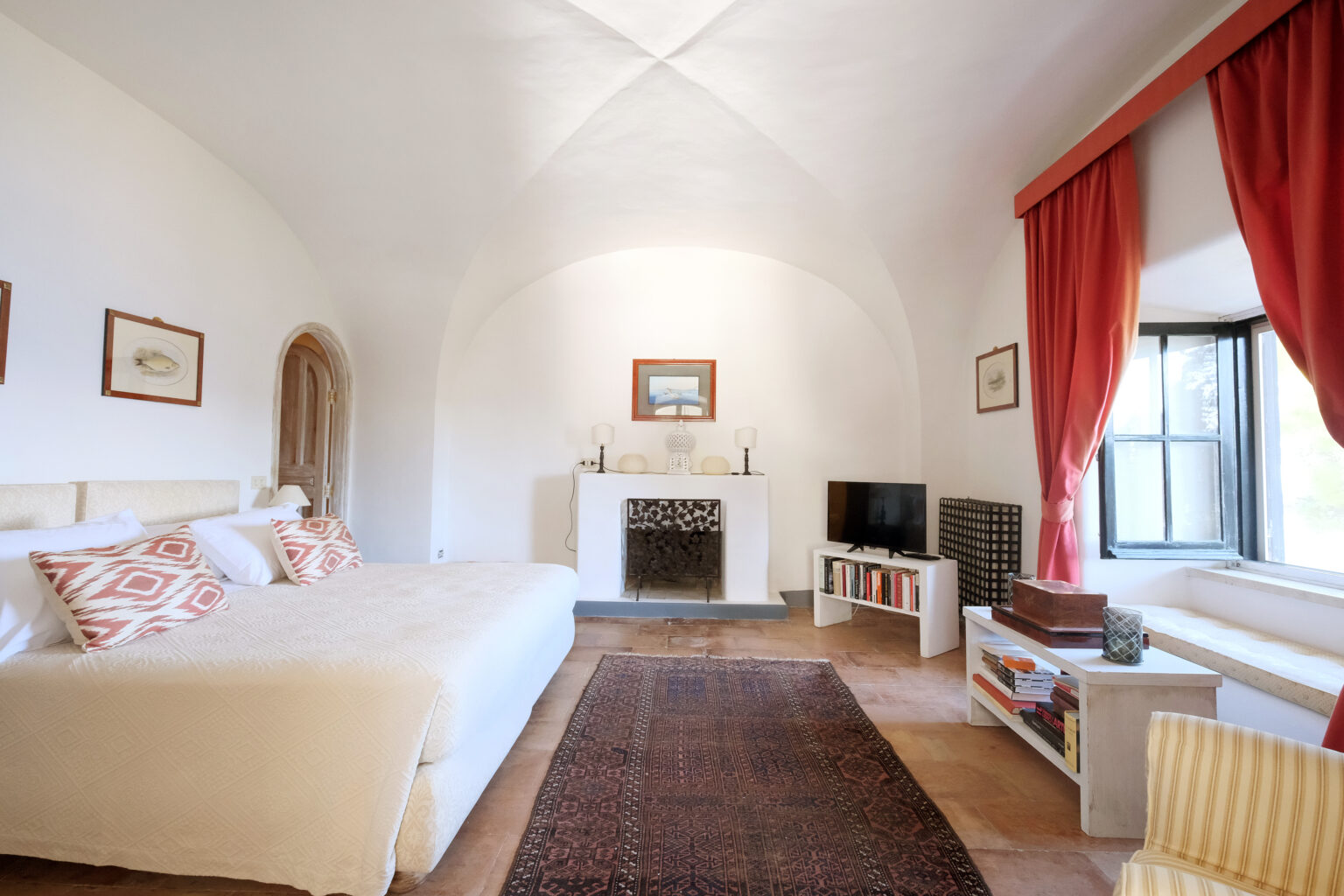 Spacious and elegantly decorated bedroom featuring a large bed, a cozy seating area, and a fireplace, complemented by warm red curtains and tasteful artwork on the walls. | Only on SopranoVillas