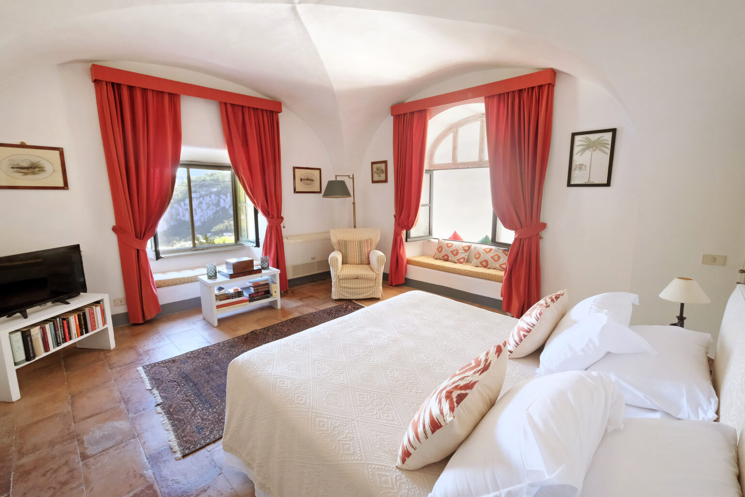 Bright and airy bedroom featuring a large bed with decorative pillows, elegant red curtains, a cozy seating area by the window, a small coffee table, and a TV unit, creating a warm and inviting atmosphere. | Only on SopranoVillas