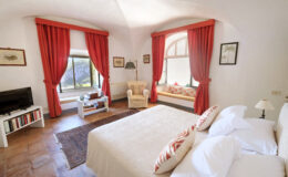 Bright and airy bedroom featuring a large bed with decorative pillows, elegant red curtains, a cozy seating area by the window, a small coffee table, and a TV unit, creating a warm and inviting atmosphere.