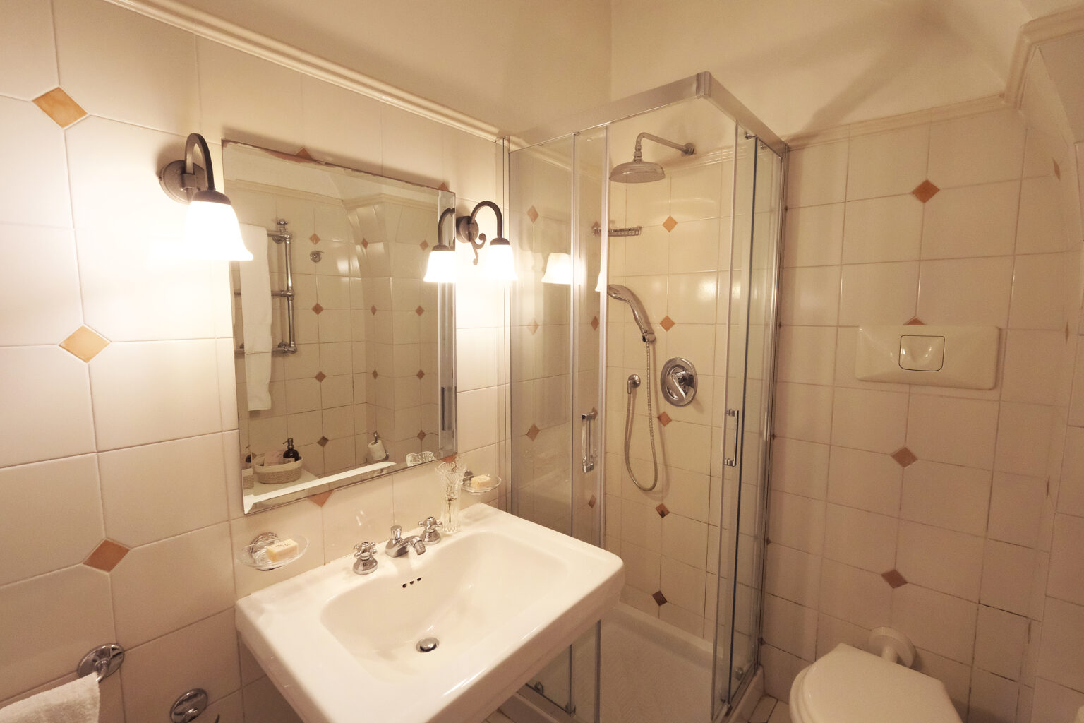 Modern bathroom featuring a glass shower enclosure, a white sink with chrome fixtures, and a large mirror, all set against tiled walls with decorative accents. Soft lighting enhances the clean and inviting atmosphere. | Only on SopranoVillas
