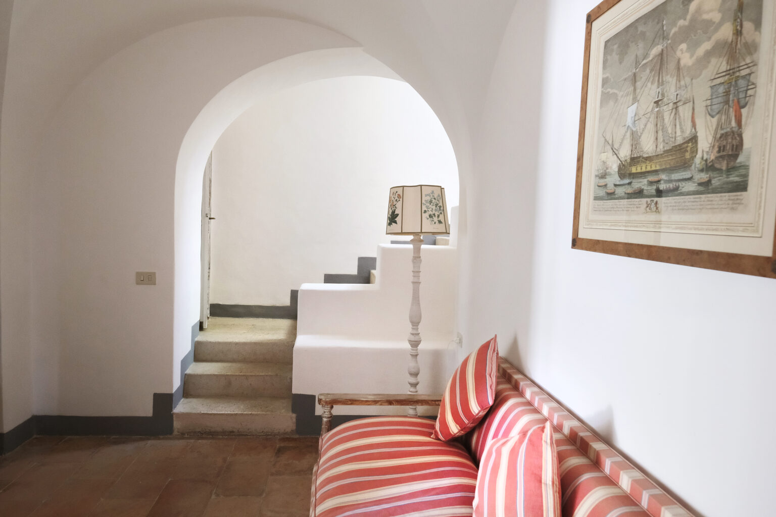 Cozy interior hallway featuring a striped sofa, a decorative lamp, and an archway leading to stairs. Artwork depicting a ship hangs on the wall, enhancing the inviting atmosphere of the space. | Only on SopranoVillas