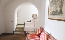 Cozy interior hallway featuring a striped sofa, a decorative lamp, and an archway leading to stairs. Artwork depicting a ship hangs on the wall, enhancing the inviting atmosphere of the space.