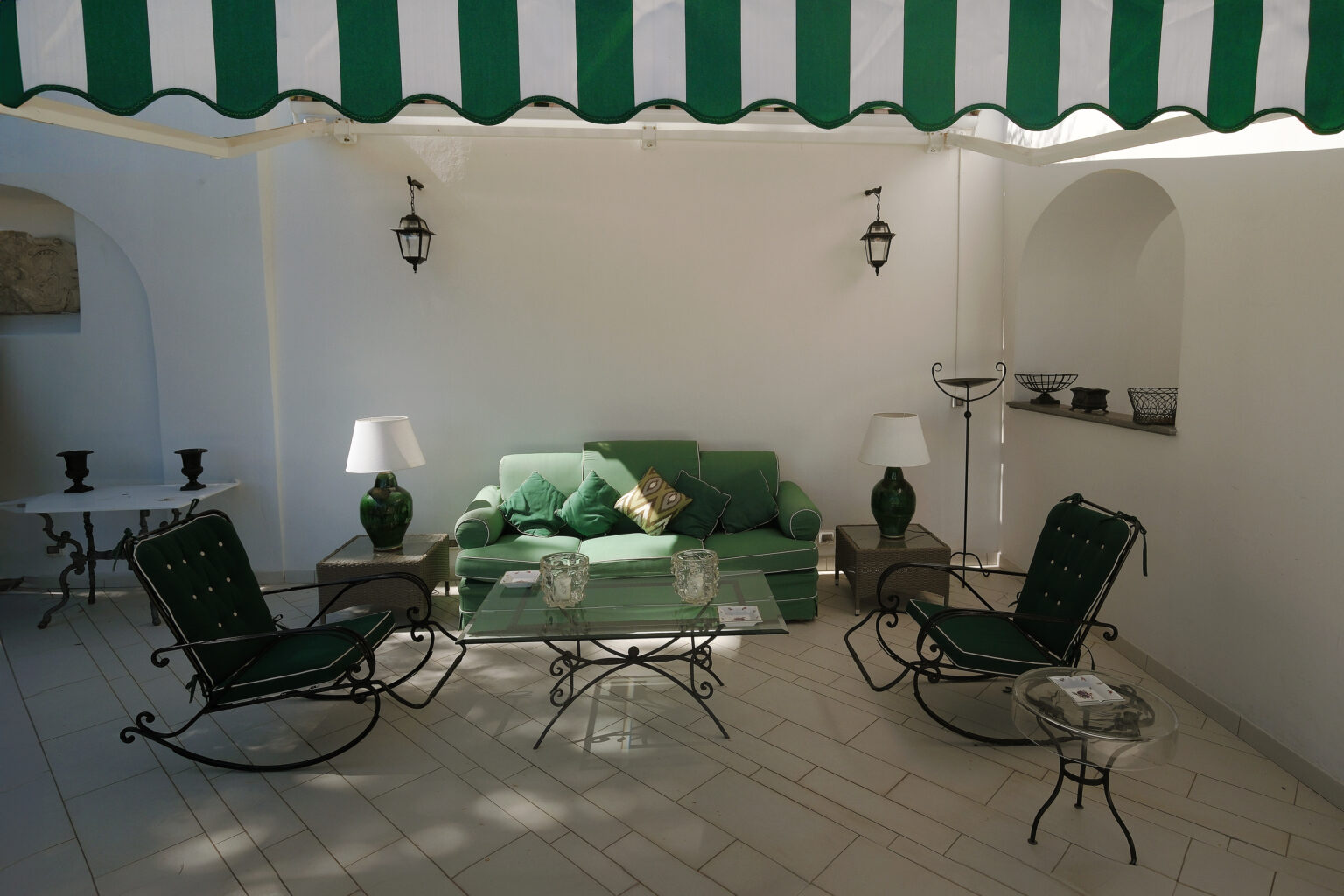 Cozy outdoor seating area featuring a green sofa, rocking chairs, and glass coffee tables, all set under a striped awning with a white wall backdrop. Ideal for relaxation and entertaining. | Only on SopranoVillas