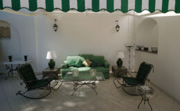Cozy outdoor seating area featuring a green sofa, rocking chairs, and glass coffee tables, all set under a striped awning with a white wall backdrop. Ideal for relaxation and entertaining.