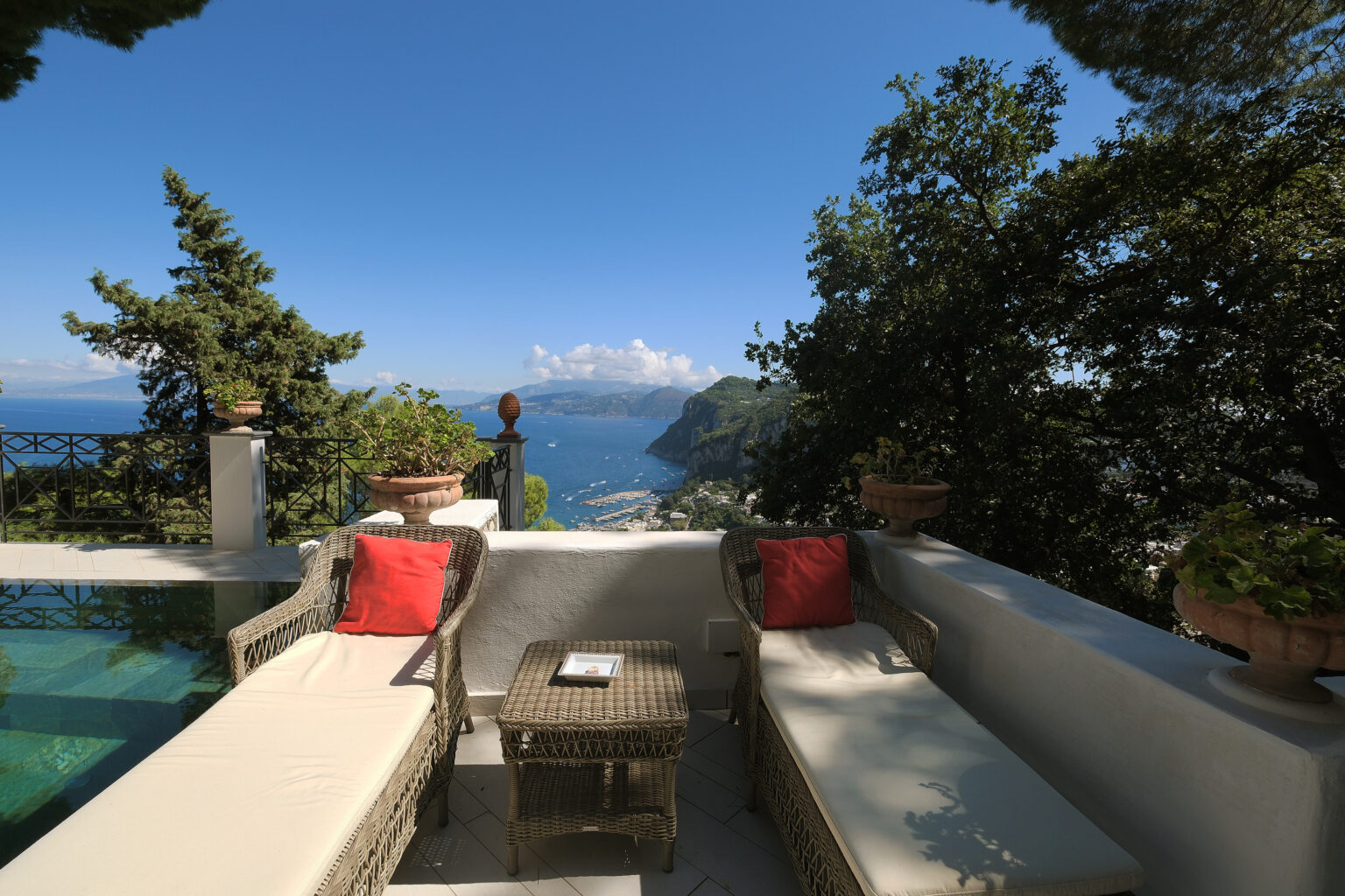 Luxurious outdoor seating area with two sun loungers and a small table overlooking a stunning coastal view, surrounded by lush greenery and a swimming pool. | Only on SopranoVillas
